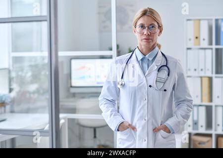 Porträt der blonden Reifen Frau als fachkundige Ärztin, die im sauberen weißen Büro steht und den Kopierraum der Kamera betrachtet Stockfoto