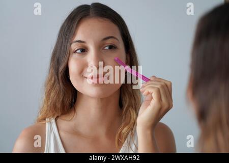 Gesichtshaarentfernung. Dermaplaning. Nahaufnahme eines schönen Mädchens, das ihr Gesicht zu Hause mit dem Rasierer rasiert. Süße junge Frau, die Rasiermesser benutzt, um Haare in der mir zu entfernen Stockfoto