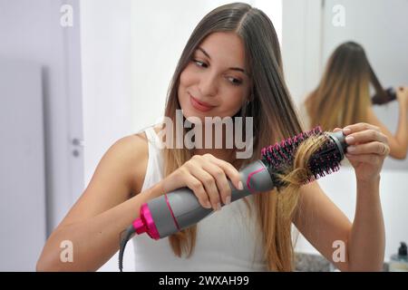 Heißluft-Haarbürste. Porträt einer jungen Frau, die zu Hause mit einer runden Bürste den Haartrockner verwendet, um die Haare auf einfache Weise zu stylen. Mädchen mit elektrischer Blowout-Bürste Haar Stockfoto
