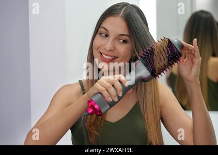 Porträt einer jungen Frau, die zu Hause mit einer runden Bürste den Haartrockner verwendet, um die Haare auf einfache Weise zu stylen. Mädchen mit elektrischer Blowout-Bürste, Haartrockner. Heißlufthaar b Stockfoto