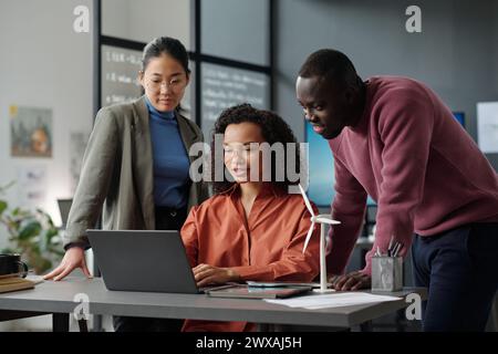 Gruppe junger, erfolgreicher interkultureller Mitarbeiter, die auf den Laptop-Bildschirm schauen, während sie Online-Daten diskutieren und ein neues Projekt vorbereiten Stockfoto