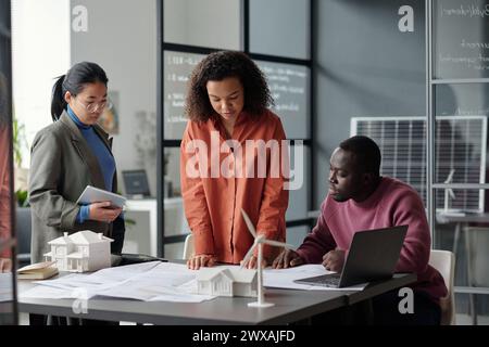Junges interkulturelles Business-Team, das sich eine Skizze auf dem Plan ansieht, während ein schwarzer Mann neben seinen weiblichen Kollegen am Schreibtisch sitzt Stockfoto