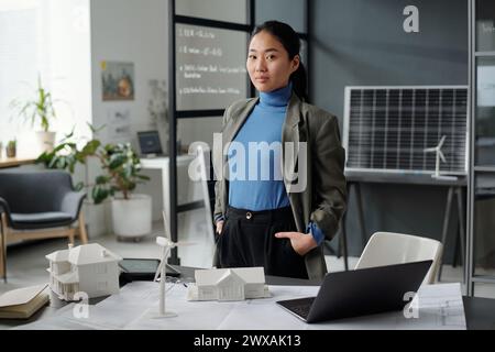 Junge zuversichtliche asiatische Geschäftsfrau in formalwear, die während der Arbeit über ein neues Architekturprojekt im Freiraumbüro in die Kamera schaut Stockfoto