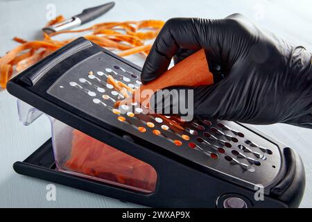 Kochen Sie in schwarzen Latexhandschuhen und zerkleinern Sie Karotten auf einer Metallreibe Stockfoto