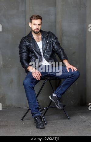 Moderner junger Mann mit stilvoller Frisur, mit einer coolen schwarzen Lederjacke und auf einem Stuhl auf dunklem Hintergrund. Stockfoto