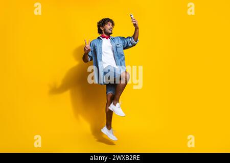 Foto in voller Länge von gutaussehendem jungen Kerl springen Selfie zeigen V-Zeichen tragen trendige Denim Kleid roter Schal isoliert auf gelbem Hintergrund Stockfoto