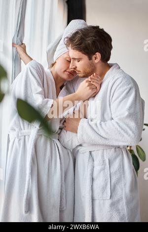 Verliebte Paare tragen Bademäntel, die sich umarmen und liebevolle Momente in der Nähe des Fensters haben Stockfoto