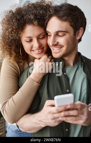 Eine lockige junge Frau küsst ihren brünetten Freund, der Kerl hält das Telefon mit einem Lächeln und geschlossenen Augen Stockfoto
