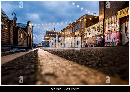 Blick auf Straßenebene entlang der urbanen Hinterstraße in der Abenddämmerung mit Graffiti und dekorativen Lichterketten. Eine Person zu Fuß Stockfoto