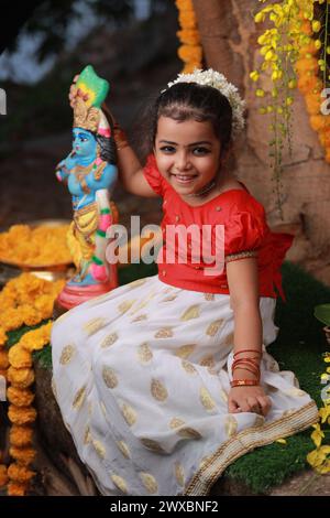 Ein niedliches kleines Mädchen, das einen langen Rock und eine rote Bluse in Kerala-Kleid trägt, sitzt unter einem Banyanbaum mit einer Statue des Herrn krishna-goldenem Sch Stockfoto