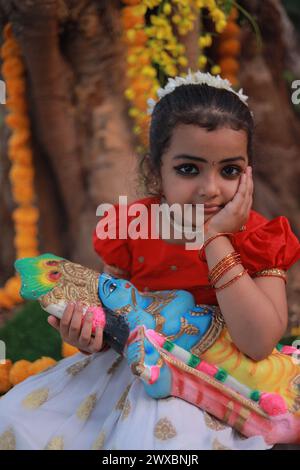 Ein niedliches kleines Mädchen, das einen langen Rock und eine rote Bluse in Kerala-Kleid trägt, sitzt unter einem Banyanbaum mit einer Statue des Herrn krishna-goldenem Sch Stockfoto