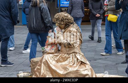 Eine lebende weibliche Statue oder eine menschliche Statue macht eine Pause, um einen McDonald's zum Mittagessen zu essen. Ein Straßenkünstler macht eine Mittagspause unter geschäftigen Einkäufern. Stockfoto