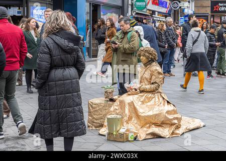 Eine lebende weibliche Statue oder eine menschliche Statue macht eine Pause, um zu Mittag zu essen, während die Menschen zusehen. Ein Straßenkünstler beobachtet die Welt beim Mittagessen Stockfoto