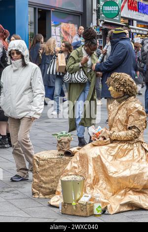 Eine lebende weibliche Statue oder eine menschliche Statue macht eine Pause, um zu Mittag zu essen, während die Menschen zusehen. Ein Straßenkünstler beobachtet die Welt beim Mittagessen Stockfoto