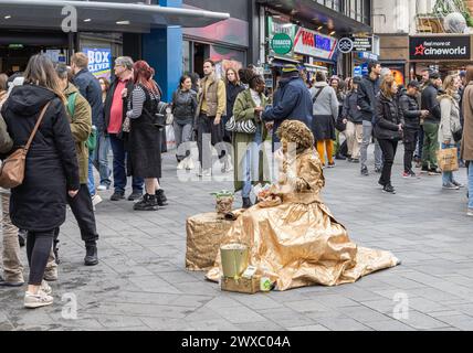 Eine lebende weibliche Statue oder eine menschliche Statue macht eine Pause, um zu Mittag zu essen, während die Menschen zusehen. Ein Straßenkünstler beobachtet die Welt beim Mittagessen Stockfoto