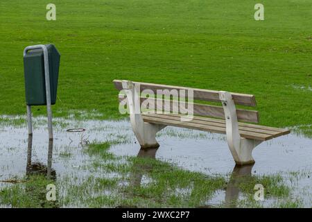 Eine öffentliche Bank und Abfalleimer im Gras, umgeben von Wasser von starkem Regen in einem Park. Stockfoto