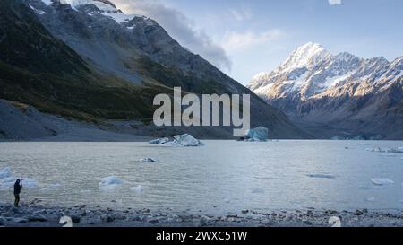 Mann, der am Ufer des Gletschersees mit Gletscher und massivem schneebedeckten Berg im Hintergrund steht. Stockfoto