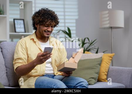 Ein entspannter Mann sitzt zu Hause auf einem Sofa mit seinem Smartphone und hält eine offene Box mit Kissen und einer Lampe in der Nähe. Stockfoto