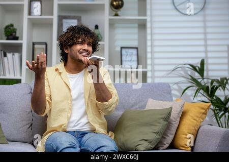 Ein junger Mann in lässigem Outfit sitzt zu Hause auf der Couch und spricht mit einem verwirrten Look auf seinem Handy, umgeben von gemütlichen Kissen und Zimmerpflanzen. Stockfoto