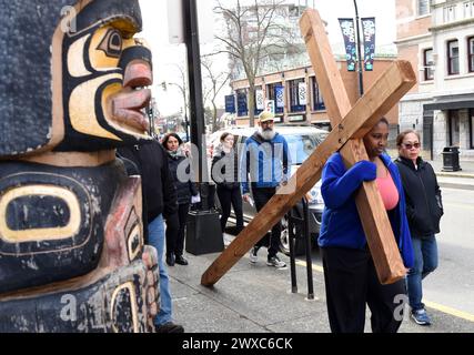 Victoria, British Columbia, Kanada, 29. März 2024 - Eine Frau trägt ein großes hölzernes Kreuz an einem Totempfahl der Indigenen in der Innenstadt von Victoria während der jährlichen Karfreitag-Passionsprozession der Christ Church Cathedral (Anglican). Don Denton/Alamy Live News Stockfoto