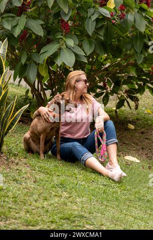 Vertikale Aufnahme einer fröhlichen Frau mittleren Alters mit ihrem Hund, beide zur Seite schauen, auf dem Gras, umgeben von Pflanzen und Blumen. Stockfoto