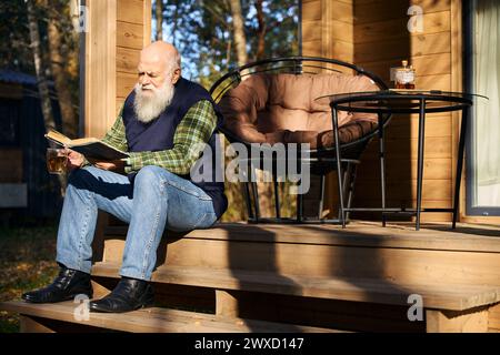 Graubärtiger Mann sitzt mit Buch auf der Veranda des Landhauses Stockfoto
