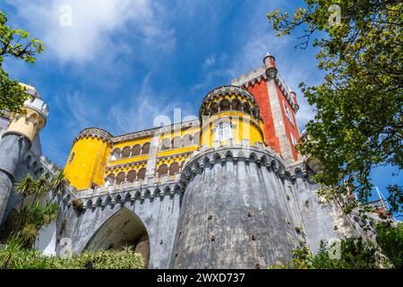 Flacher Blick auf die befestigten Mauern des Pena Palace in einem bunten gelb-roten Stil an Wänden, die mit einigen Palmen unter einem sonnigen blauen s bedeckt sind Stockfoto