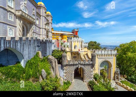 Außenansicht und breiter Blick auf den farbenfrohen Pena Palast mit dem Weg hinauf zum bogenförmigen Eingang, der mit Basreliefs verziert ist, unter einem sonnigen blauen Himmel. Sintra. Stockfoto