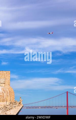 Westliches Profil des Monuments für die Entdeckungen, mit Touristen auf dem Dachboden, die auf die Brücke 25 de Abril blicken und ein kommerzielles Flugzeug durch die Brücke fliegen Stockfoto