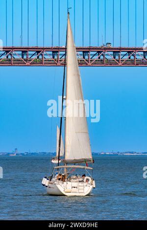 Zwei touristische Erholungssegelboote segeln auf dem Tejo mit der roten Stahlbrücke 25 de Abril darüber mit Auto- und Lastkraftverkehr und Stockfoto