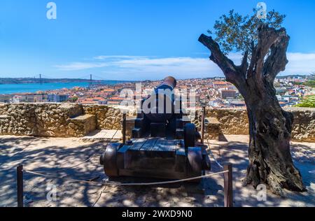 Restaurierte Bronzekanone von St. George's Castle mit Blick auf die Stadt Lissabon und die rote Metall 25 de Abril Hängebrücke neben einem Olivenbaum. Stockfoto