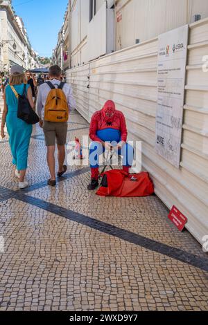 Fetter übergewichtiger Mann, gekleidet als spiderman auf einer Straße in Lissabon, sitzend und auf sein Handy schauend, verkaufte Figuren von fetten Spinnen mit Touristen Pass Stockfoto