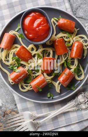 Festliche lustige Essensspaghetti mit Würstchen und Ketchup Nahaufnahme auf einem Teller auf dem Tisch. Vertikale Draufsicht von oben Stockfoto