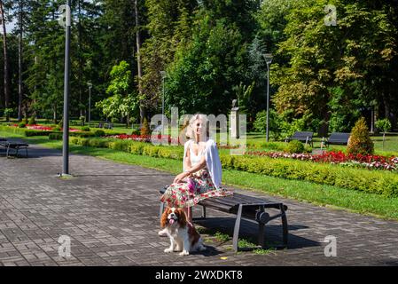 Frau und Hund - Kavalier King Charles Spaniel - Entspannen in einem öffentlichen Park mit üppigen Blumen, Pflanzen, Gras und Bäumen Stockfoto
