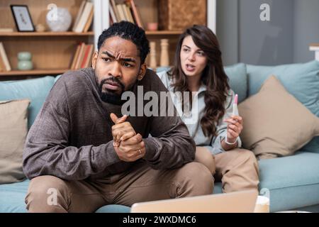 Unglückliche junge Frau, die auf dem Sofa sitzt, mit Schwangerschaftstest und afroamerikanischem Ehemann, drinnen. Millennials-Familie im Konflikt. Stockfoto