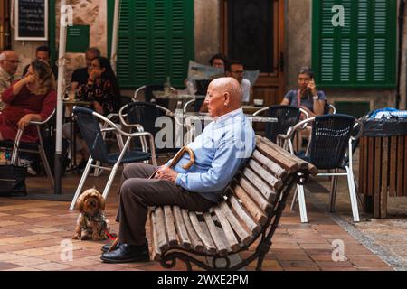 Fornalutx, Mallorca, Spanien - 7. Mai 2023: Älterer Mann mit Hund ruht auf einer Bank in einem typischen Bergdorf in der Nähe eines Cafés. Kontrast von friedlich Stockfoto