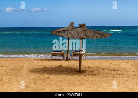 Ein leerer Strand in der Nebensaison mit Strohschirmen und blauer Sonnenliege. Playa del Castillo , Fuerteventura, Kanarische Inseln, Spanien Stockfoto