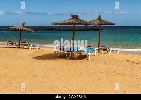 Ein leerer Strand in der Nebensaison mit Strohschirmen und blauer Sonnenliege. Playa del Castillo , Fuerteventura, Kanarische Inseln, Spanien Stockfoto