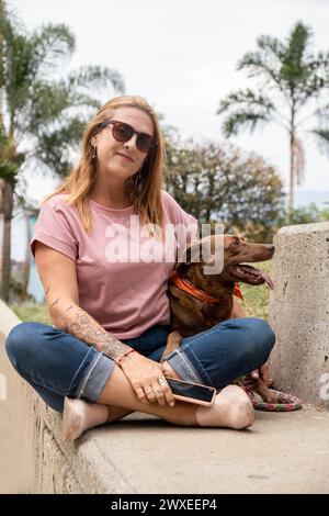 Frau mittleren Alters mit Sonnenbrille, sitzt auf einer Zementbank neben ihrem Hund, mit Palmen im Hintergrund. Stockfoto