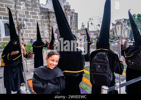 Antigua, Guatemala. März 2024. Ein junges Mädchen lächelt, als sie mit einer Brüderschaft von Bußbürgern posiert, während sie vor der Kirche Escuela de Cristo auf den Beginn der Karfreitagsprozession des Senor Sepultado während der Semana Santa, 29. März 2024 in Antigua, Guatemala, warten. Die opulente Prozession ist eine der größten der Welt mit Tausenden von Gläubigen und dauert 12 Stunden. Quelle: Richard Ellis/Richard Ellis/Alamy Live News Stockfoto