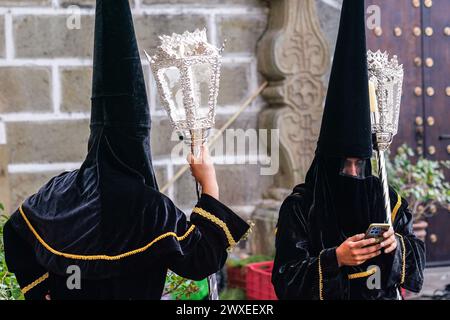 Antigua, Guatemala. März 2024. Eine Brüderschaft von Pönitenten, die Capirot-Hüte tragen, überprüft ihre Handys, während sie vor der Kirche Escuela de Cristo auf den Beginn der Karfreitagsprozession des Senors Sepultado während der Semana Santa, 29. März 2024 in Antigua, Guatemala, warten. Die opulente Prozession ist eine der größten der Welt mit Tausenden von Gläubigen und dauert 12 Stunden. Quelle: Richard Ellis/Richard Ellis/Alamy Live News Stockfoto