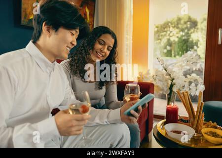 Zwei lächelnde Freunde trinken Wein und schauen während der Feiertagsfeier zu Hause telefonisch Stockfoto