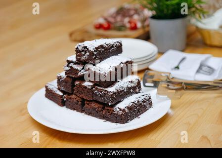 Frisch gebackene Schokoladen-Brownies, mit Puderzucker bestäubt, auf einem weißen Teller gestapelt, mit Geschirr im Hintergrund. Stockfoto