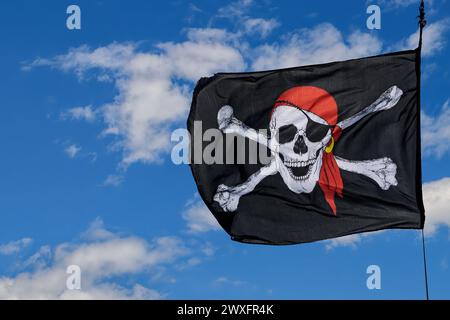 Eine schwarze, weiße und rote generische Jolly Roger schlägt in den Winden gegen einen teilweise bewölkten blauen Himmel. Der Schädel hat einen Augenfleck und einen goldenen Ohrring. Stockfoto