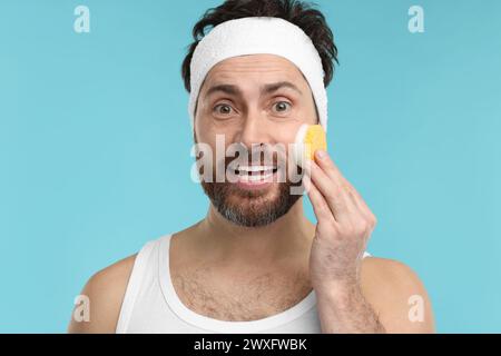 Mann mit Stirnband, der sein Gesicht mit Schwamm auf hellblauem Hintergrund wäscht Stockfoto