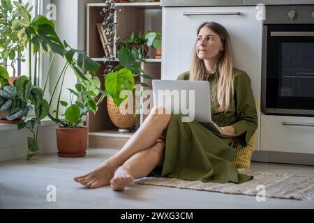 Nachdenkliche Frau mit Laptop, die auf das Fenster blickt, Gefühl von Energiemangel, sitzen zu Hause mit Pflanzen auf dem Boden Stockfoto