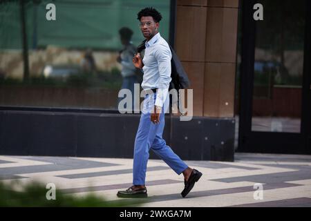 Stilvoller Afroamerikaner spaziert in der Stadt draußen, trägt blaue Hosen und ein Hemd mit Loafers und strahlt urbanen Raffinesse aus. Stockfoto