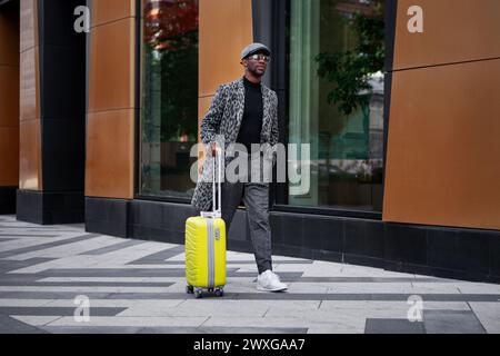 Stilvoll gekleidetes männliches Model geht die Stadtstraße mit gelbem Koffer in der Hand. Schwarzer Mann Tourist draußen in der Nähe des Hotelgebäudes. afroamerikaner Stockfoto