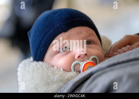 Extreme Nahaufnahme eines glücklichen kleinen Babys mit großen Augen, die mit einem Schnuller und einem blauen Hut im Freien nach oben schauen Stockfoto