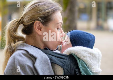 SideView einer attraktiven blonden Mutter in ihren 40ern, die ihr neugeborenes Baby liebevoll in einer Babytrage hält und ihm sanft auf die Nase küsst Stockfoto